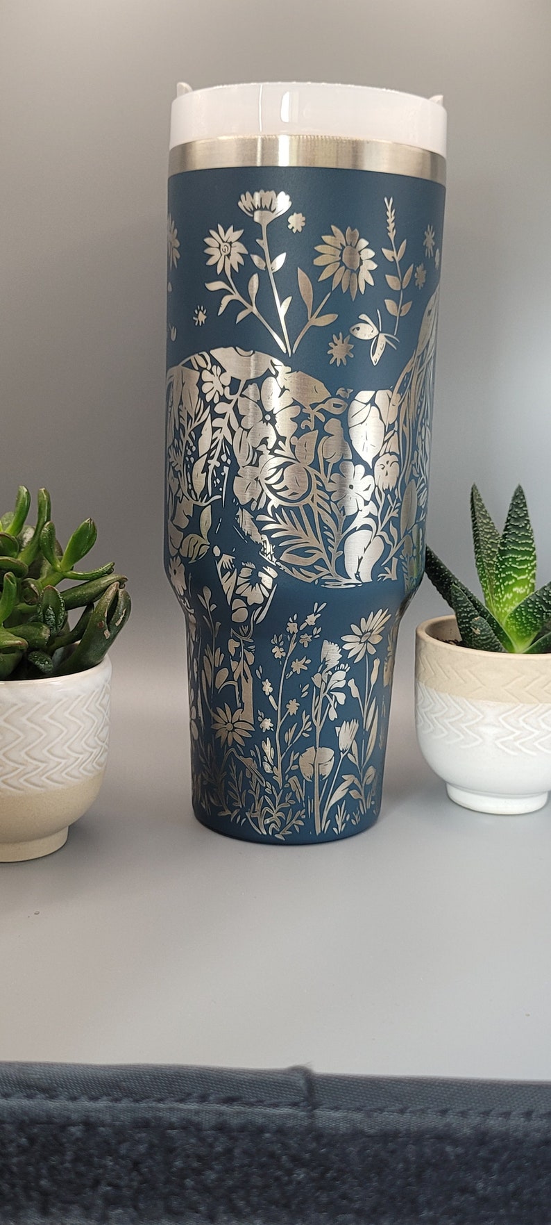 May include: A blue stainless steel tumbler with a silver floral design. The tumbler has a white lid and is perfect for keeping drinks cold or hot.