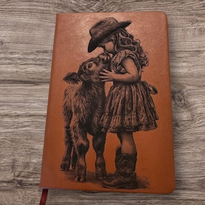 Op de afbeelding: Een bruin leren notitieboek met een illustratie van een jong persoon met een cowboyhoed die een kalf kust. De zwart-wit kunst is geplaatst tegen een warme, oranje-bruine achtergrond. Het notitieboek heeft een rood lint als bladwijzer.