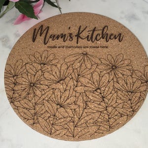Puede incluir: Posavasos redondo de corcho con el texto "Mum's Kitchen" y "meals and memories are made here". El posavasos presenta un diseño floral negro. El fondo es de color corcho natural.