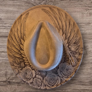May include: A tan fedora hat with a decorative brim featuring a detailed wing design and rose pattern. The hat has a teardrop crown and a decorative cord. The hat is a neutral colour and would be suitable for many outfits.