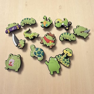 Puede incluir: Una colección de dijes de zapatos verdes de estilo caricatura con varios animales y objetos. Los diseños incluyen ranas, una tortuga, una serpiente, un lagarto y un cocodrilo. Algunos dijes tienen accesorios.