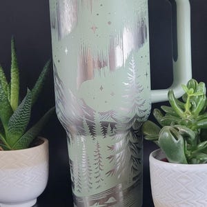 May include: A mint green insulated tumbler with a handle, featuring a silver etched design of a mountain range, trees, and stars. The tumbler has a white lid and a matching green handle. Two small potted plants flank the tumbler.