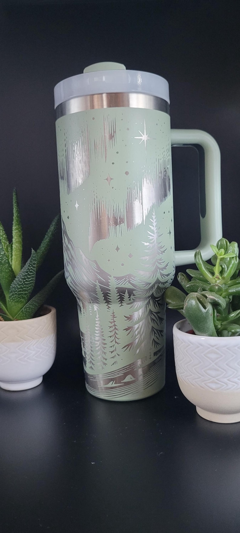 May include: A mint green insulated tumbler with a handle, featuring a silver etched design of a mountain range, trees, and stars. The tumbler has a white lid and a matching green handle. Two small potted plants flank the tumbler.