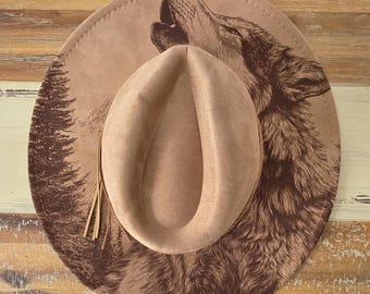 May include: A tan cowboy hat with a detailed illustration of a howling wolf and a forest scene. The hat features a brown suede crown and a decorative tassel. The hat's brim measures approximately 15 inches in diameter.