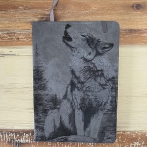 Op de afbeelding: Grijze notitieboek met een gedetailleerde illustratie van een huilende wolf in een bos. De kaft heeft een textuur en een grijs lint als bladwijzer. De wolf staat centraal, met bomen op de achtergrond.