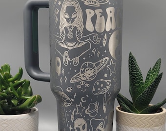 May include: A gray stainless steel travel mug with a black handle. The mug is decorated with a white design of a meditating alien, mushrooms, planets, stars, and the word "Peace".