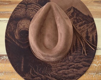May include: A brown wide-brimmed hat featuring a dark brown laser-engraved design on the brim. The artwork depicts a large bear face on the left side and a mountain landscape with pine trees on the right. A braided band and thin fringe accent the crown.