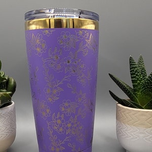 Op de afbeelding: Een paarse roestvrijstalen beker met een gouden deksel en een bloemenpatroon. Het patroon bestaat uit gouden bloemen en bladeren op een paarse achtergrond.