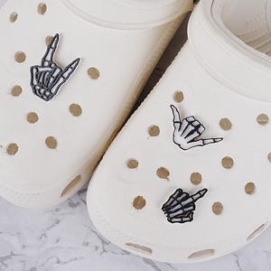 May include: A pair of off-white slip-on clogs featuring circular ventilation holes. The left clog is decorated with a black and white skeleton hand charm making a rock-and-roll gesture, while the right clog features two skeleton hand charms.