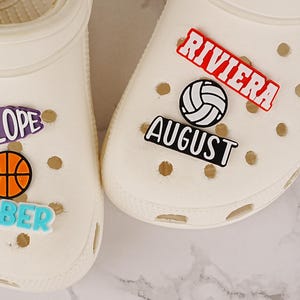 Peut inclure: Deux sabots en mousse blanc cassé décorés de breloques colorées. Le sabot gauche porte les inscriptions PENELOPE et TIMBER avec un ballon de basket. Le sabot droit porte les inscriptions RIVIERA et AUGUST avec un ballon de volley.