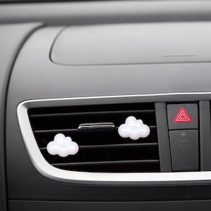 Könnte beinhalten: Fünf weiße, wolkenförmige Autolufterfrischer, die an den Lüftungsschlitzen im Armaturenbrett eines Autos hängen. Die Lufterfrischer bestehen aus einem weichen, biegsamen Material und haben eine leicht strukturierte Oberfläche.