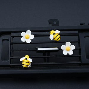 Puede incluir: Cuatro clips de ambientador de coche con un diseño de abeja amarilla y negra y un diseño de margarita blanca. Los clips están unidos a una rejilla de ventilación de coche negra.