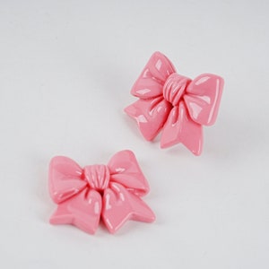 Puede incluir: Un par de pendientes de lazo de plástico rosa. Los lazos son brillantes y tienen un efecto 3D.