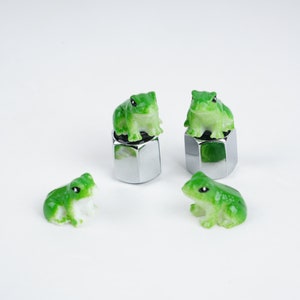 May include: Four green frog figurines, three of which are perched on silver metal caps. The frogs have black eyes and white bellies.