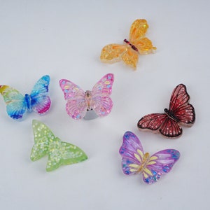 Könnte beinhalten: Sechs bunte Schmetterlings-Anhänger mit schimmernden Glitzer-Akzenten. Die Schmetterlinge sind in den Farben Pink, Blau, Grün, Gelb, Lila und Rot.