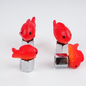 May include: Four red plastic goldfish valve stem caps for car tires. The caps are attached to silver metal valve stems.