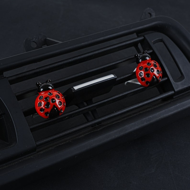 May include: Two red and black ladybug car air freshener clips. The ladybugs are attached to a black car air vent.