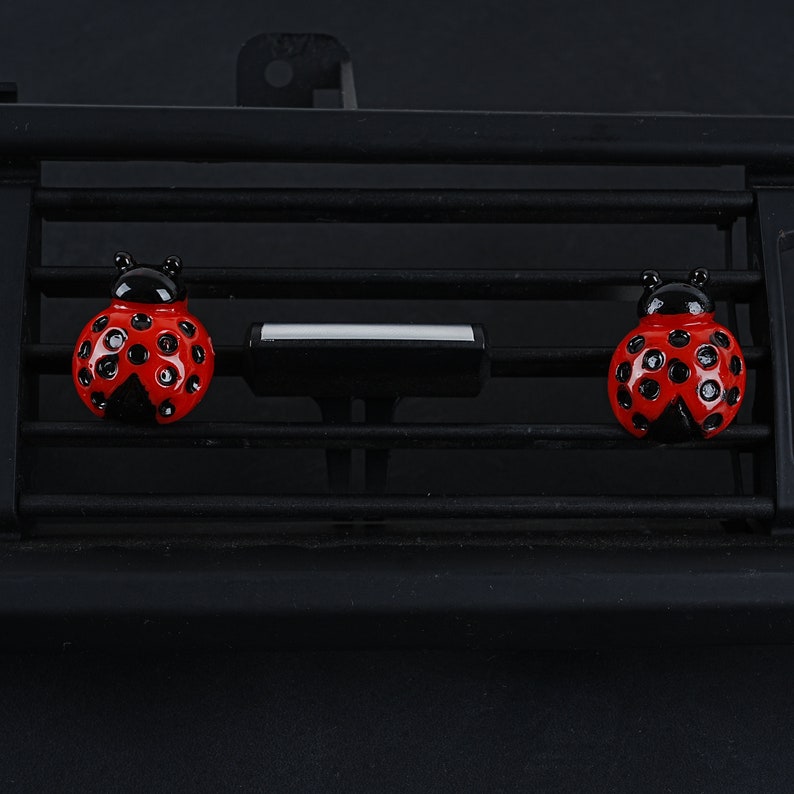 May include: Three red ladybug car air fresheners with black spots and black heads are attached to a black car air vent.