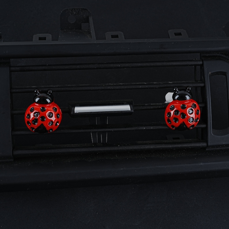 May include: Two red and black ladybug car air fresheners hanging from a black car air vent.