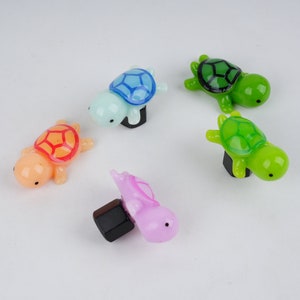 May include: Five colorful turtle figurines with black eyes and painted shells. The turtles are made of glass and have a shiny finish. The turtles are arranged in a circle on a white background.