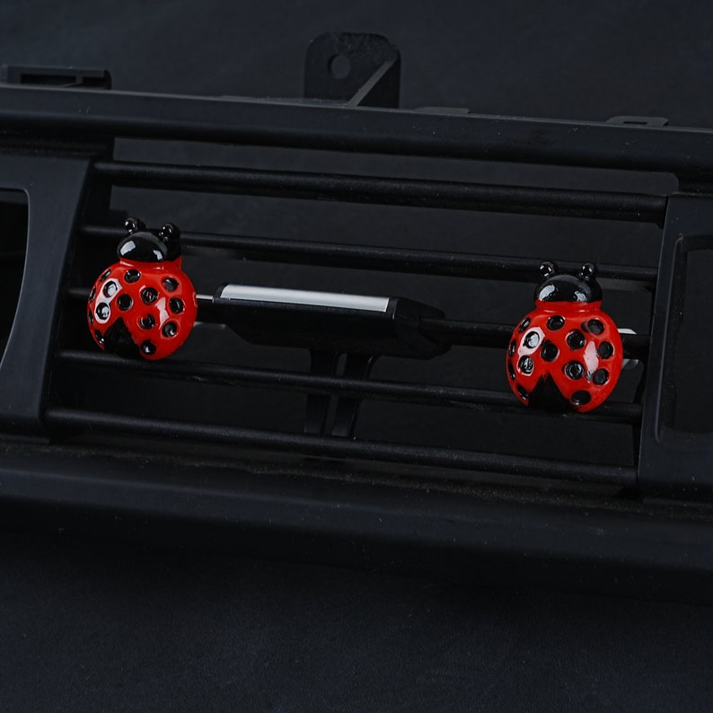 May include: Two red ladybug car air freshener clips with black spots and antennae. The clips are attached to a black car air vent.
