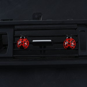 May include: Two red ladybug car air fresheners with black spots and black heads are attached to a black car air vent.