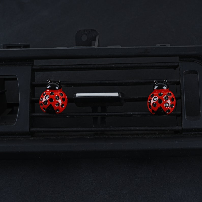 May include: Two red ladybug car air fresheners with black spots and black heads are attached to a black car air vent.
