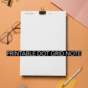 Op de afbeelding: Een wit bedrukbaar stippenraster notitiepapier met de tekst "NOTES" en "plan your work, work your plan" erop gedrukt. Het papier is vastgeklemd aan een perzikkleurig oppervlak met een gouden paperclip. Er liggen gouden paperclips verspreid over het oppervlak.