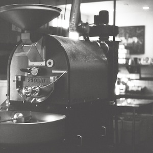 Könnte beinhalten: Ein schwarzer und silberner Kaffeeröster mit einem großen Trichter und einer Trommel. Der Röster ist mit "Probat" beschriftet und hat einen silbernen Griff.