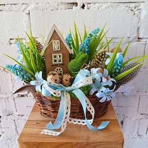 Puede incluir: Una cesta de mimbre llena de una casa de madera en miniatura, flores azules, musgo verde y huevos de codorniz. La cesta está decorada con una cinta azul y encaje blanco.