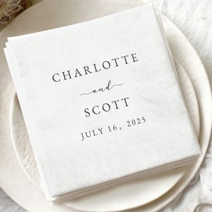 May include: A pile of white paper napkins printed with the names "Charlotte and Scott" and the date "July 16, 2025" in black. The napkins are on a white plate, with two rings in the foreground.