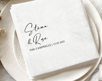 May include: A stack of white paper napkins with the names "Steven & Rose" and the date "15.05.2025" printed in black. The napkins are on a white plate, with two wedding rings in the bottom right corner.