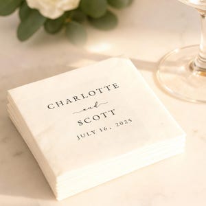 May include: A stack of white paper napkins with the names "Charlotte and Scott" and the date "July 16, 2025" printed in black. The napkins are stacked neatly, suggesting a wedding or event. A glass and greenery are in the background.