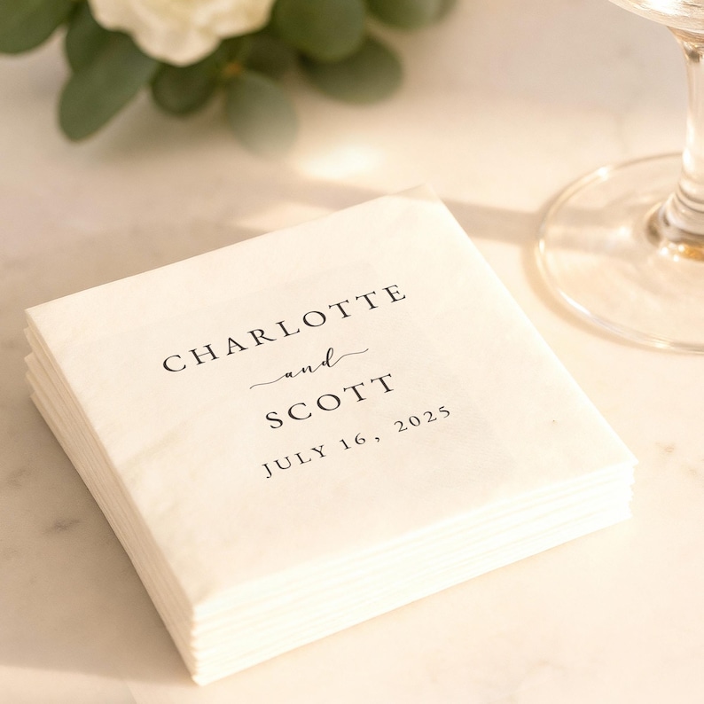 May include: A stack of white paper napkins with the names "Charlotte and Scott" and the date "July 16, 2025" printed in black. The napkins are stacked neatly, suggesting a wedding or event. A glass and greenery are in the background.