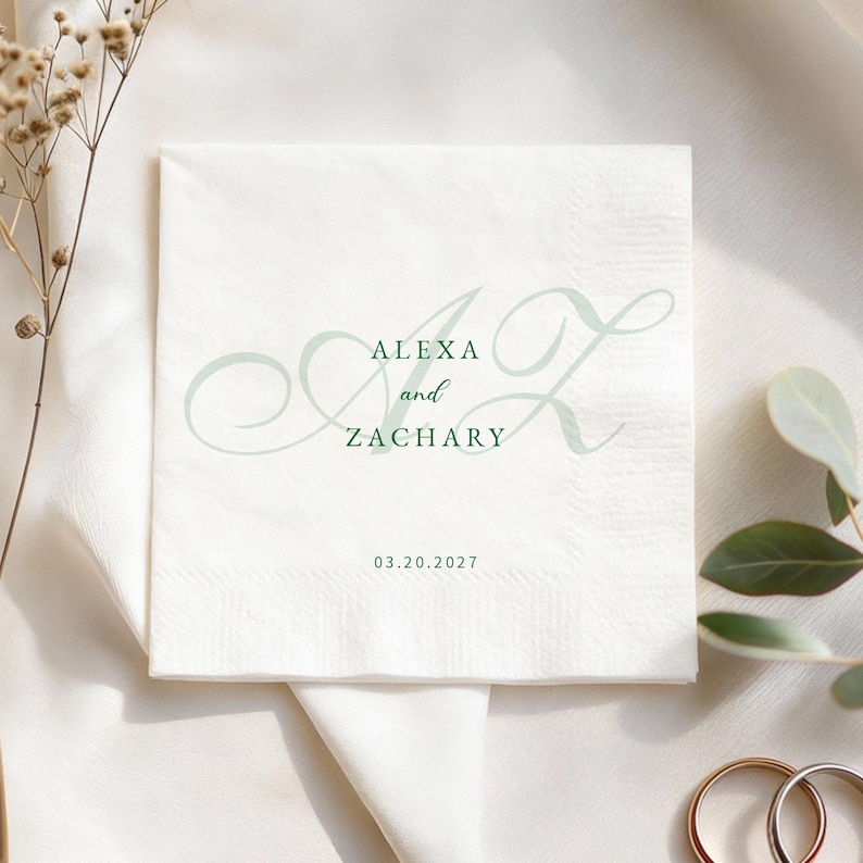 May include: White square paper napkin with the names "Alexa and Zachary" and the date "03.20.2027" printed in a soft green script. The napkin is folded and sits on a white surface, with decorative elements in the background.