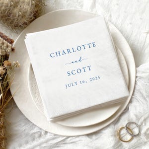 May include: A stack of white paper napkins with the names "Charlotte" and "Scott" printed in navy blue, along with the date "July 16, 2025". The napkins are on a white plate, with wedding rings in the foreground.