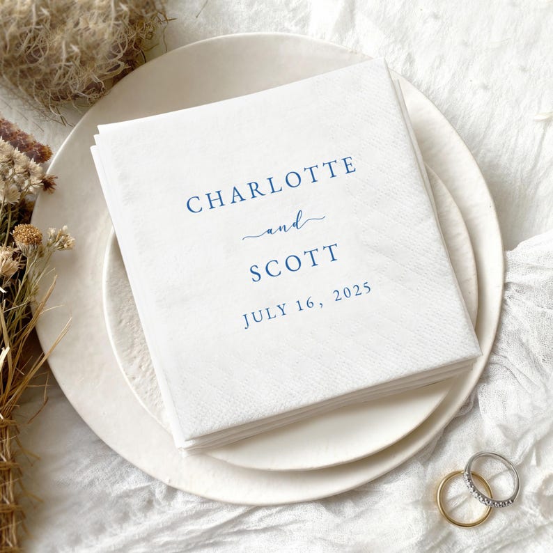May include: A stack of white paper napkins with the names "Charlotte" and "Scott" printed in navy blue, along with the date "July 16, 2025". The napkins are on a white plate, with wedding rings in the foreground.