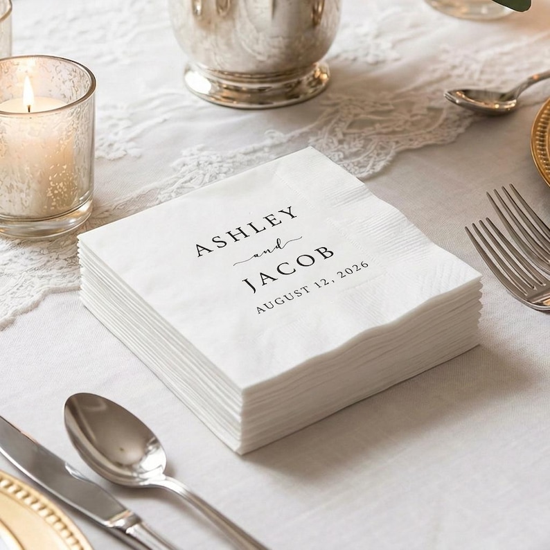 May include: A stack of white paper napkins with the names "ASHLEY and JACOB" and the date "AUGUST 12, 2026" printed in black. The napkins are on a white lace tablecloth, with a lit candle and silverware in the background.