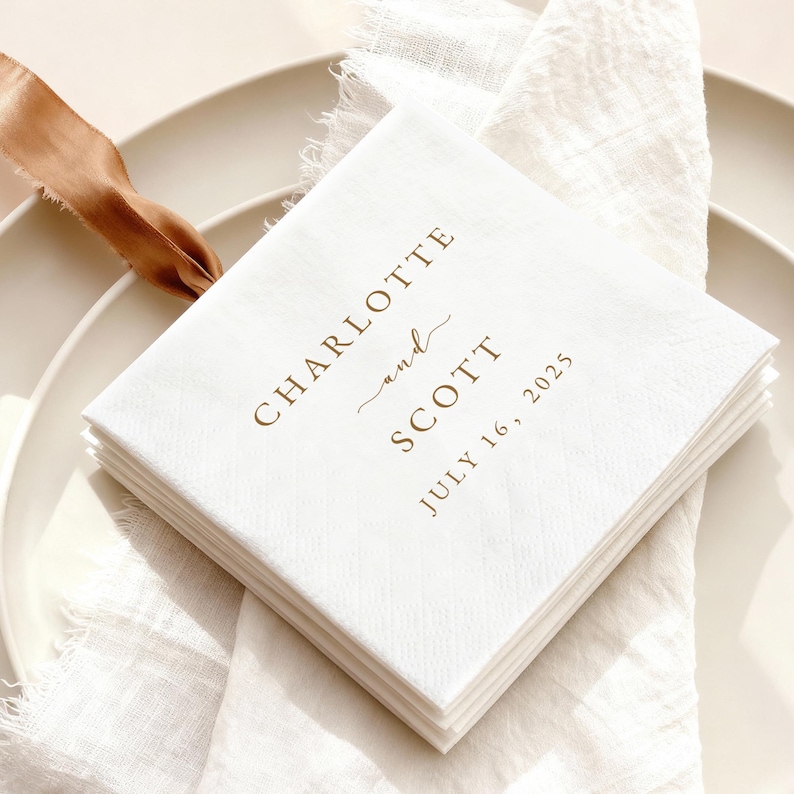 May include: A stack of white square napkins with the names "CHARLOTTE and SCOTT" and the date "JULY 16, 2025" printed in brown. The napkins are on a white plate with a white cloth and a brown ribbon.