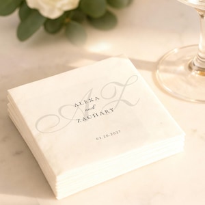 May include: A stack of white paper napkins with the names "Alexa and Zachary" and the date "03.20.2021" printed in gray script. The napkins are stacked on a light-colored surface, with a glass and greenery in the background.