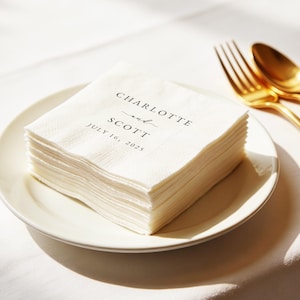 May include: A stack of white paper napkins on a white plate. The napkins are printed with the names "Charlotte and Scott" and the date "July 16, 2025" in black script. Gold fork and spoon are visible.