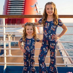 May include: Two young girls wearing matching navy blue jumpsuits with a Minnie Mouse print. The jumpsuits have short sleeves and flared legs. The background shows a cruise ship and the ocean. The clothing is from the brand "ran clothing kids".