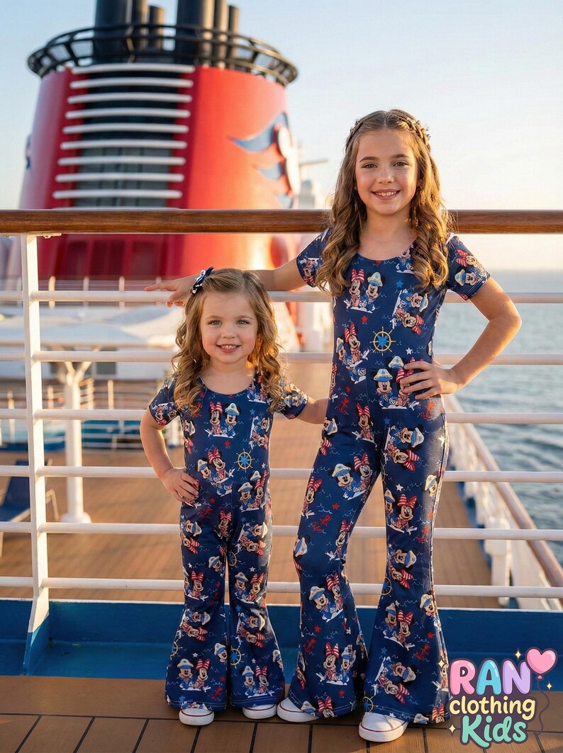 May include: Two young girls wearing matching navy blue jumpsuits with a Minnie Mouse print. The jumpsuits have short sleeves and flared legs. The background shows a cruise ship and the ocean. The clothing is from the brand "ran clothing kids".