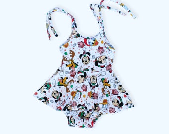 May include: A white baby onesie with a Christmas-themed print featuring Mickey Mouse, Minnie Mouse, Donald Duck, and Goofy. The onesie has a ruffled skirt and adjustable straps.