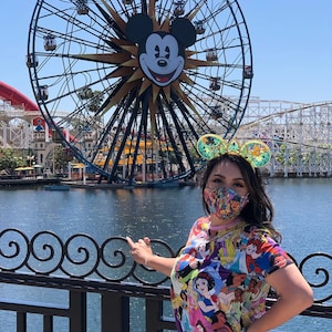 May include: A woman wearing a colorful Disney princess t-shirt and a pair of Mickey Mouse ears stands in front of a Ferris wheel with Mickey Mouse's face on it. The Ferris wheel is in the background, and the woman is in the foreground. She is wearing a face mask.