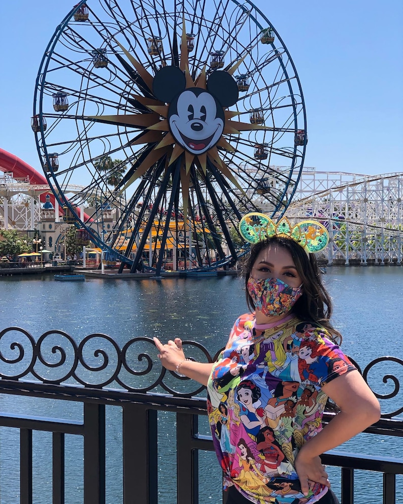 May include: A woman wearing a colorful Disney princess t-shirt and a pair of Mickey Mouse ears stands in front of a Ferris wheel with Mickey Mouse's face on it. The Ferris wheel is in the background, and the woman is in the foreground. She is wearing a face mask.