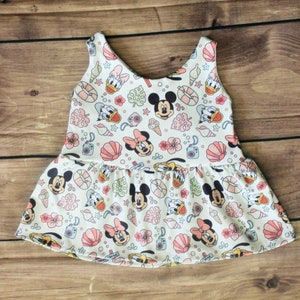 May include: A white sleeveless dress with a pink, green, and blue print featuring Mickey Mouse and Donald Duck characters. The dress has a ruffled skirt and a scoop neck.