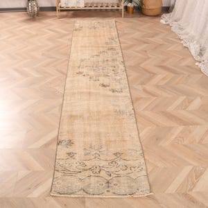 May include: A beige and cream runner rug with a faded floral pattern. The rug has a distressed look and is made of a soft, woven material.