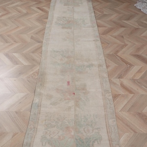 May include: A long, rectangular rug with a faded, vintage appearance. The rug features a cream-colored background with floral patterns in shades of green, pink, and blue. The edges are bordered with a darker trim. The rug is placed on a wooden floor.