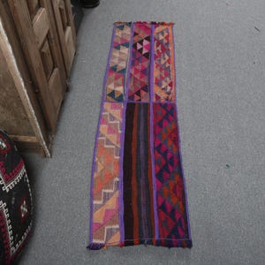 May include: A long, rectangular runner rug featuring a patchwork design of various woven textile panels. The rug displays a vibrant color palette of purple, orange, red, and black, with geometric patterns including triangles and zigzags.